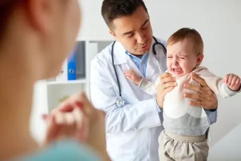 Lekarz uspokaja płaczące dziecko, którego rodzice sprawdzają skoki rozwojowe w tabeli.